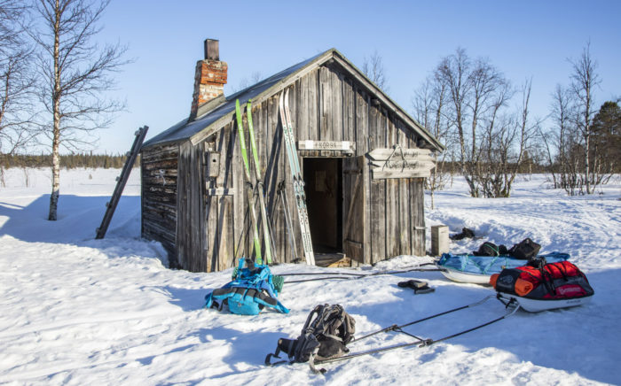 Skis and sleds are placed in front of a small wooden building surrounded by a snowy landscape.