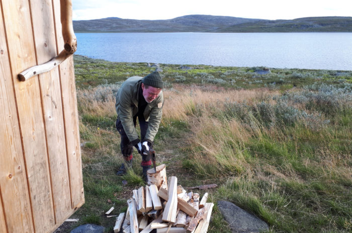 A man chops wood with a lake and mountains in the background.