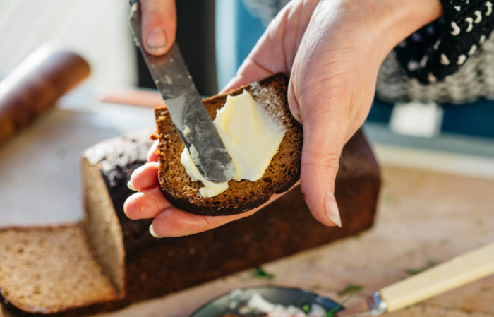 Las manos de una persona mientras extienden una capa de mantequilla sobre una rebanada de pan negro.