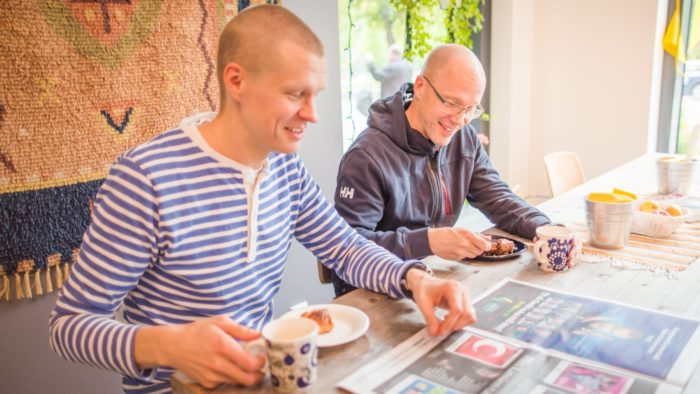At a café table, two men are reading a newspaper, drinking coffee and eating buns.