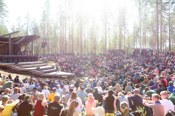Debout sur une scène aménagée en plein air avec une forêt visible à l’arrière-plan, un homme tient un discours à un public composé de plusieurs centaines de jeunes gens.