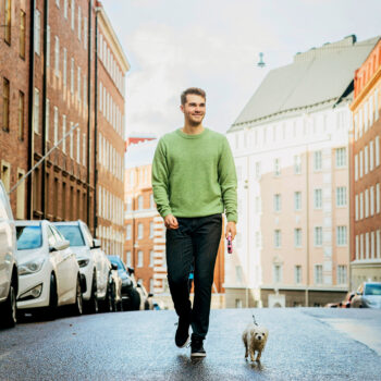 رجل مبتسم يمشي مع كلب صغير في شارع في مدينة هلسنكي خلال يوم مشمس.