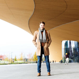 Ein Mann in Mantel und Schal steht vor einem Gebäude aus Holz und Glas.