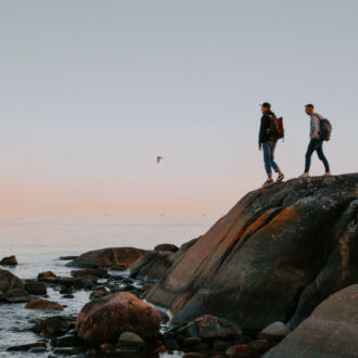 Two people look out over the sea from the top of a large boulder on a rocky shoreline.