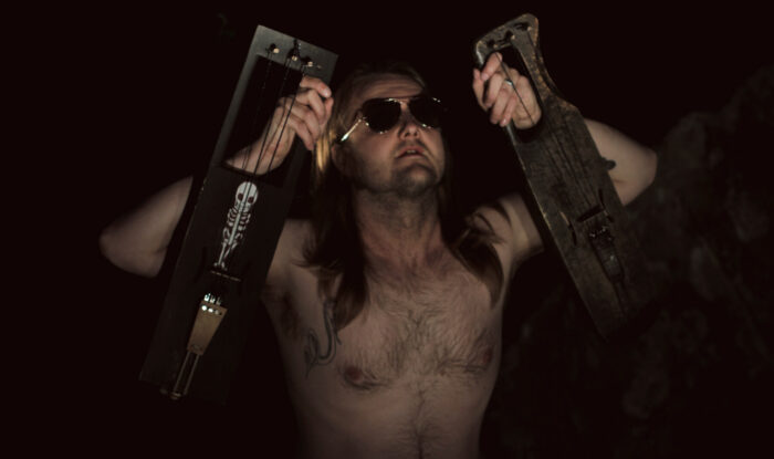Мужчина с голым торсом и в темных очках держит в обеих руках по деревянному струнному инструменту.