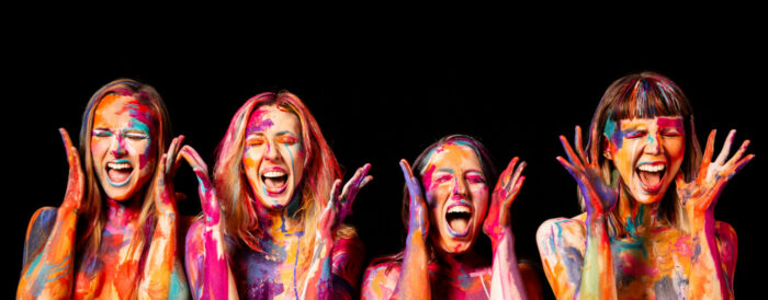 Vier Frauen, deren Arme und Gesichter mit bunten Farbklecksen bedeckt sind, stehen in einer Reihe und singen mit weit geöffneten Mündern.