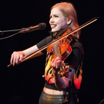 A woman plays a violin while singing into a microphone.