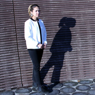 A woman in a white jacket and black trousers stands in front of a wooden wall, casting a shadow on the wall.
