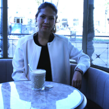 Uma mulher com uma jaqueta branca está sentada em frente a uma janela em uma cafeteria com uma xícara de café.