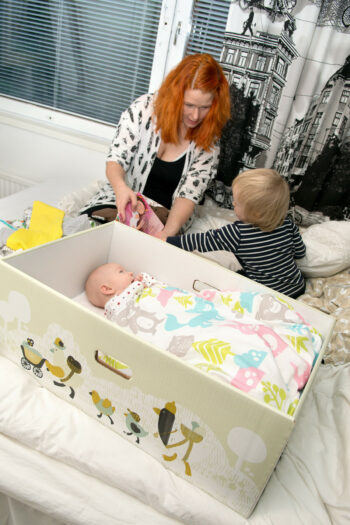 Eine Frau und ein Kleinkind sitzen neben einem Karton, in dem ein Baby unter einer bunten Decke schläft.