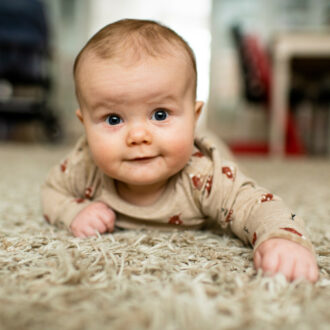 A smiling baby in a light brown onesie crawling on a light brown carpet.