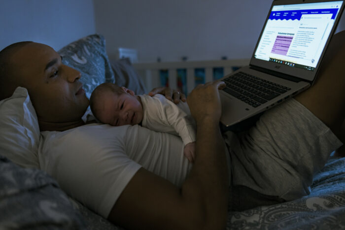 Ein Mann liegt auf einem Bett mit einem Baby auf der Brust und einem Laptop auf den angewinkelten Beinen.