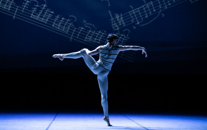 A lone dancer balances on one foot in front of a picture of music notes.