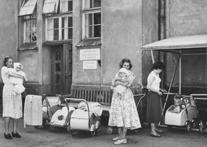 Uma foto em preto e branco de várias mulheres segurando bebês ao lado de uma fileira de carrinhos em frente a um prédio.
