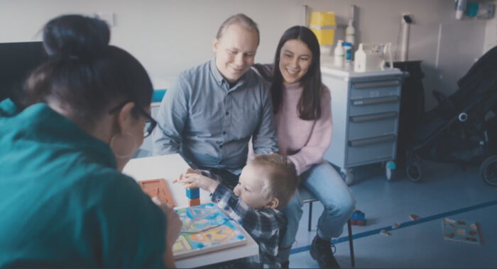 A young child arranges building blocks on a table while his parents and a nurse watch him.