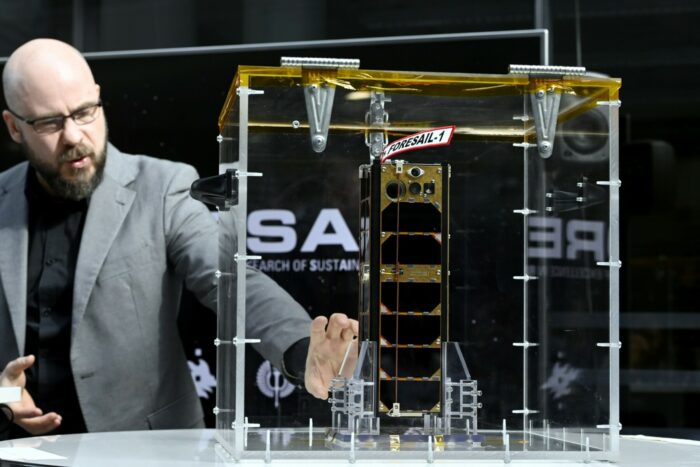A man gestures towards a rectangular metal construction that is on display in a glass case.