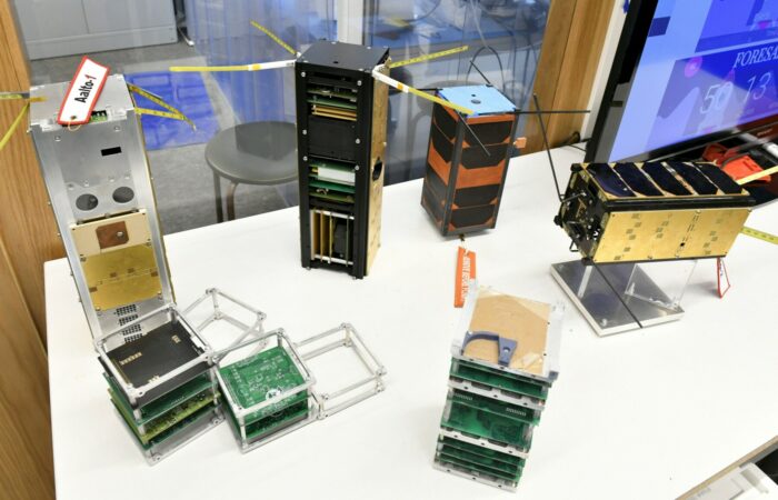 Several satellites that look like rectangular metal boxes are on display on a table.