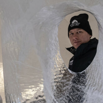 A man dressed in winter clothing looks at the camera through a hole in an ice sculpture.
