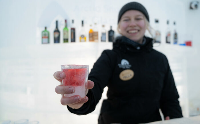 A bartender in winter clothing holds up a glass made of ice filled with red liquid.