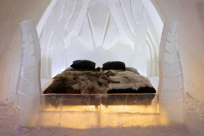 Blankets and pillows are arranged on a bed-size block of ice in a room made entirely of ice and snow.