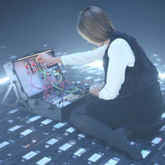 Sobre el suelo de un escenario cubierto de casetes de VHS, una mujer con vestido negro y camisa blanca está sentada ante una mesa de mezclas de sonido.