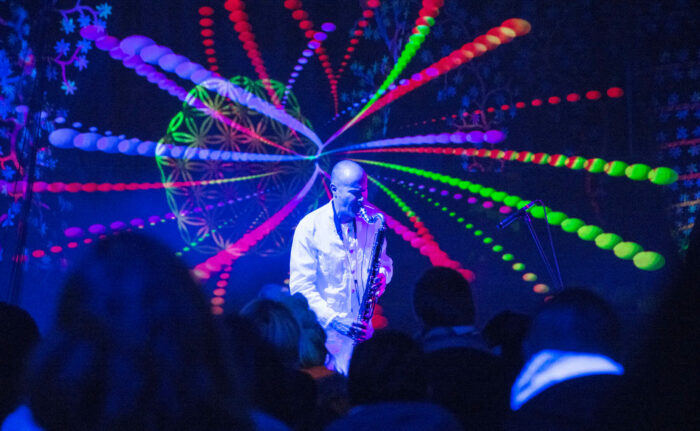 Un hombre vestido de blanco toca un clarinete bajo en un escenario con coloridos efectos de luces a sus espaldas.