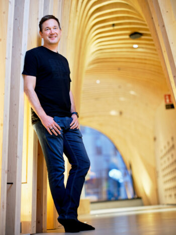 Um homem de jeans e camiseta preta está encostado na parede de um corredor com arcos de madeira.