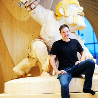 A man in blue jeans and a black T-shirt sits in front of a wooden statue of a bearded warrior.