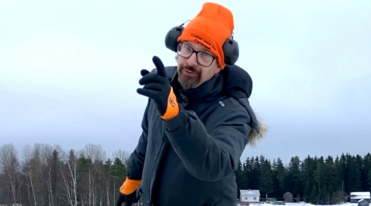 A man dressed in heavy winter clothing holds up one finger and smiles.