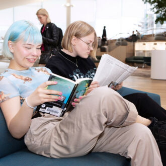 Two people read on a sofa in a large, open library space while others browse books or talk with each other in the background.