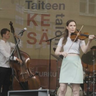 A woman plays the violin on a summery open-air stage, accompanied by three men playing keyboards, double bass and drums.