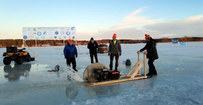 A man pushes a circular saw mounted on a wooden frame along a frozen lake surface while several people watch.