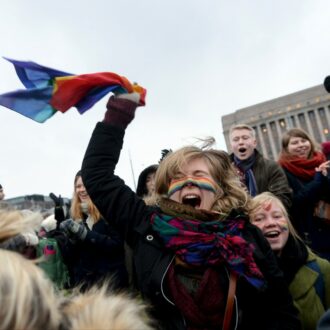 群众呼喊着，挥舞着彩虹色旗帜，背景中可见议会大厦。