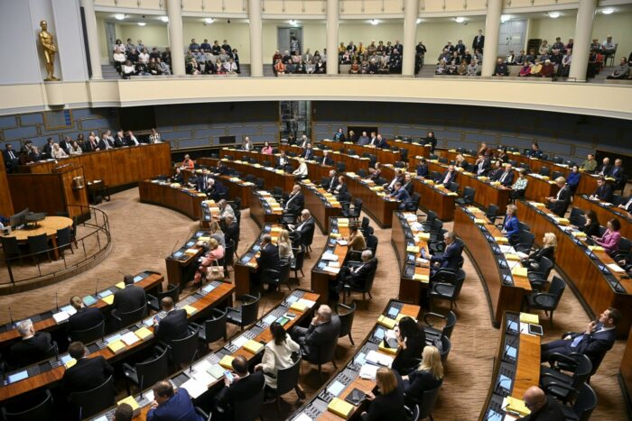 议会议员坐在大厅里，他们面前的桌子排列成一个个向心半圆形状，旁听的群众则成排坐在楼座的椅子上。