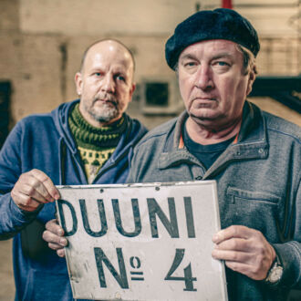 Deux hommes vêtus de bleus de travail tiennent entre leurs mains une pancarte sur laquelle figure un court texte écrit en caractères rétro.