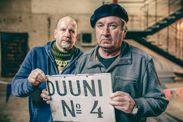 Deux hommes vêtus de bleus de travail tiennent entre leurs mains une pancarte sur laquelle figure un court texte écrit en caractères rétro. 