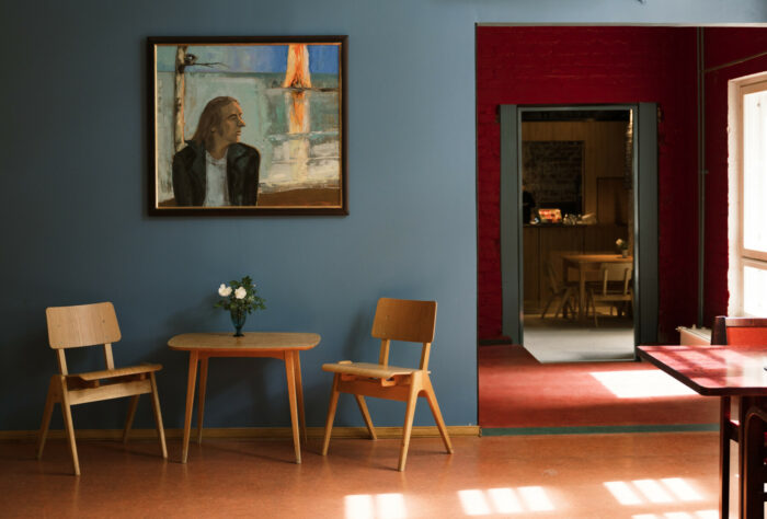 A luz do sol entra pelas janelas em uma sala onde uma pintura de uma cena de verão está pendurada na parede acima de uma mesa e algumas cadeiras.