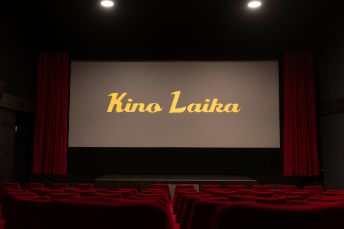 The interior of a cinema contains rows of plush red seats and a large screen flanked by red curtains.