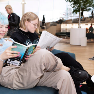 Deux personnes sont installées sur un canapé dans une vaste salle de bibliothèque tandis que d’autres visiteurs feuillettent des livres ou échangent entre eux à l’arrière-plan.