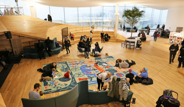 Varios adultos y niños descansan y juegan en una gran sala de techo abovedado, luminosa y llena de alfombras de colores.