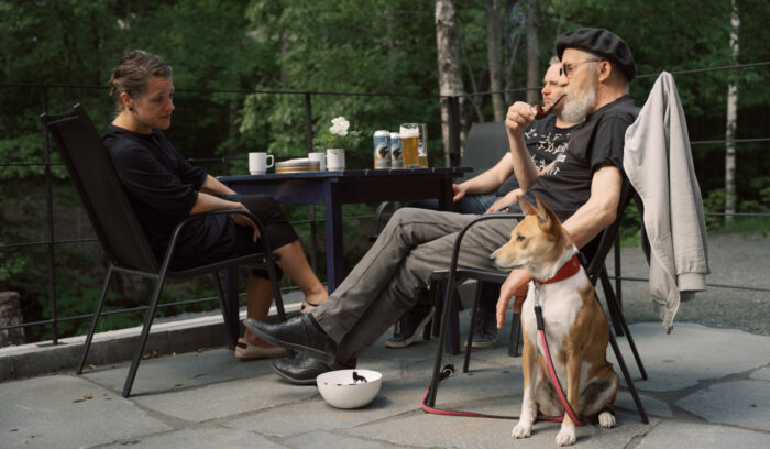 Una mujer y dos hombres sentados alrededor de una mesa al aire libre, con un perro junto a ellos. 
