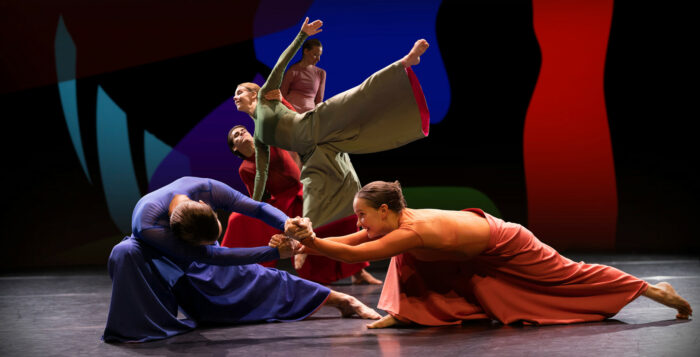 Cinq ou six danseurs et danseuses habillés de costumes de scène de couleurs claires se tiennent en équilibre dans des poses variées. 