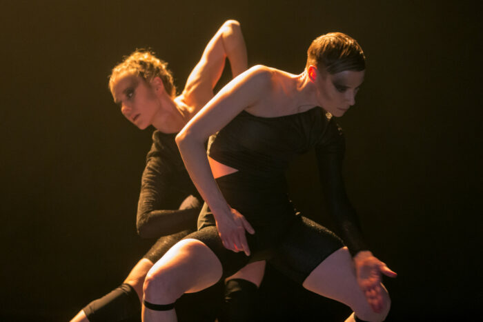 In costumes made of bands of black fabric, two dancers stand together while bending in opposite directions.
