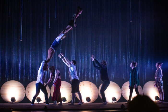 A tower of three acrobats standing on each other’s shoulders is leaning and falling, as other people wait to catch them.