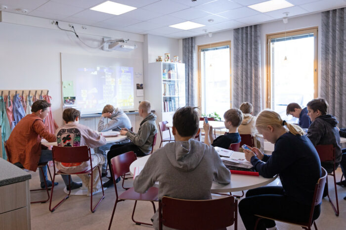 Schüler, die in einem Klassenzimmer an runden Tischen sitzen, schauen auf mobile Geräte, die sie in der Hand halten.