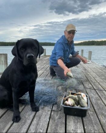 Чёрная собака и мужчина на пирсе на фоне озера. На первом плане фотографии миска с рыбой и рыболовная сеть.