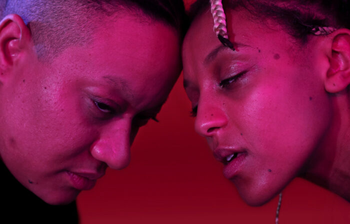 Num close-up que parece banhado por uma luz rosa, duas pessoas encostam a testa uma na outra.