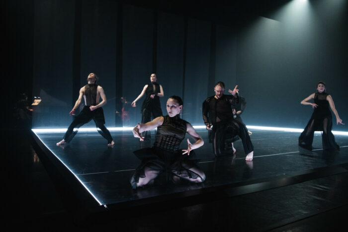 Auf einer Bühne mit einem Beleuchtungsband auf Bodenhöhe tanzen fünf schwarz gekleidete Tänzer.