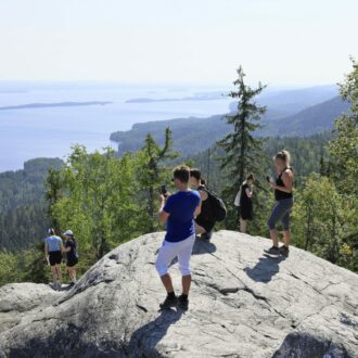 Several people in T-shirts and shorts look out over a vast lake from a large rock on a forested ridge.