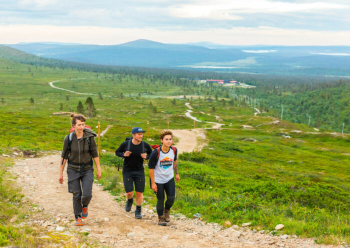 Três jovens com mochilas sobem uma colina com picos de montanhas visíveis ao fundo.